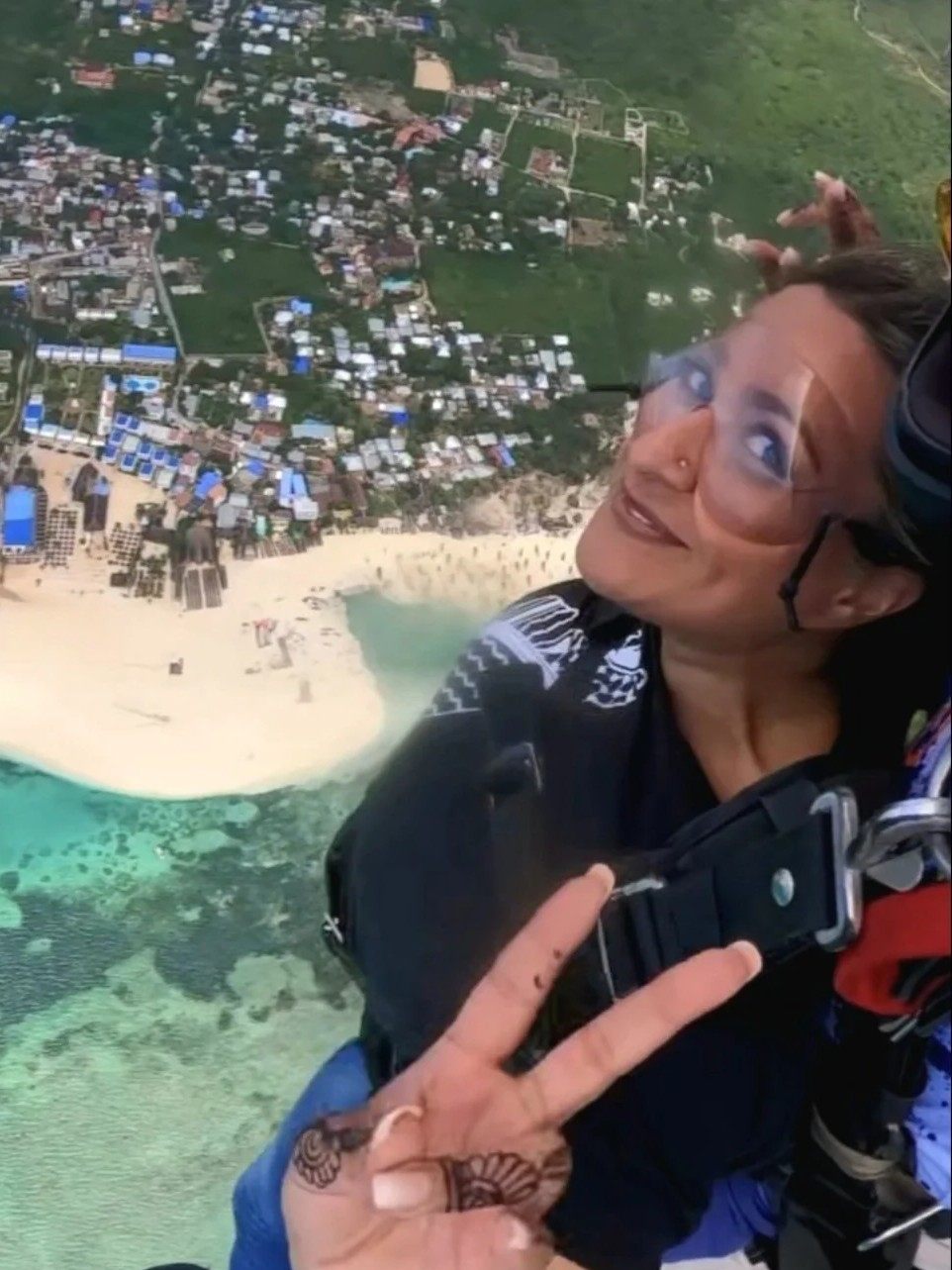 Anira Khokhar tandem skydiving above a coastline, wearing goggles and a harness and making a peace sign