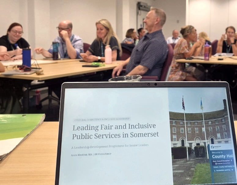 A laptop showing the title slide of Anira Khokhar's programme “Leading Fair and Inclusive Public Services in Somerset”, with participants seated at tables behind