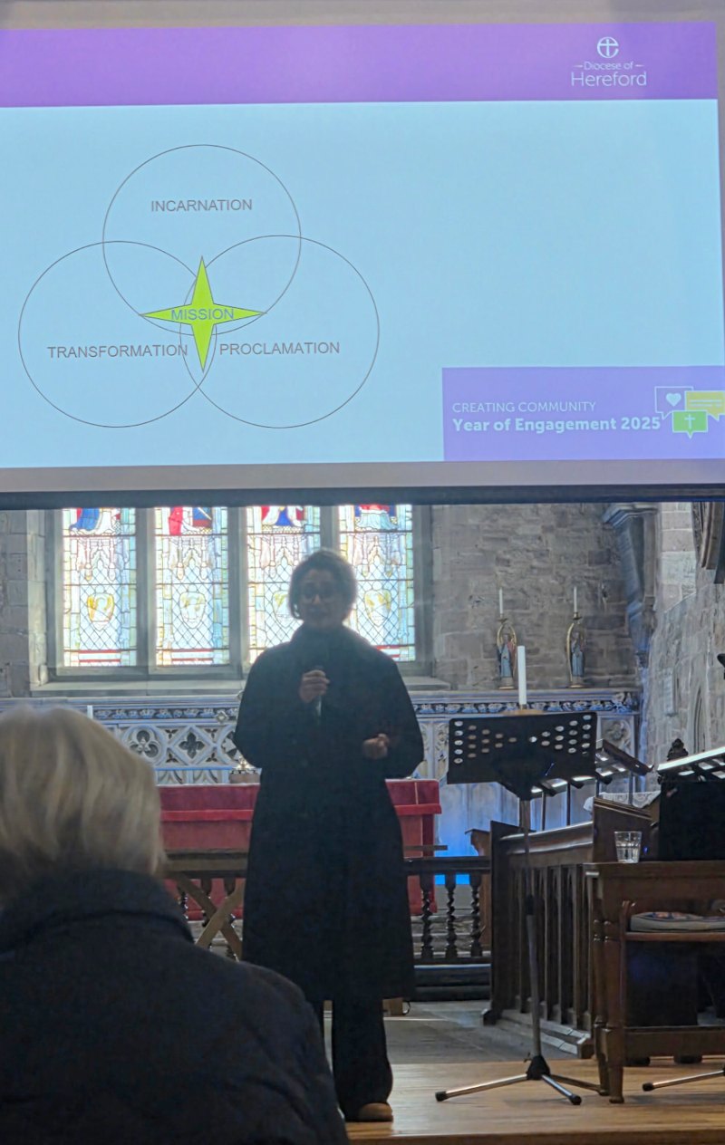 Anira Khokhar speaking into a microphone in a church, beneath a Diocese of Hereford slide reading “Creating Community, Year of Engagement 2025”