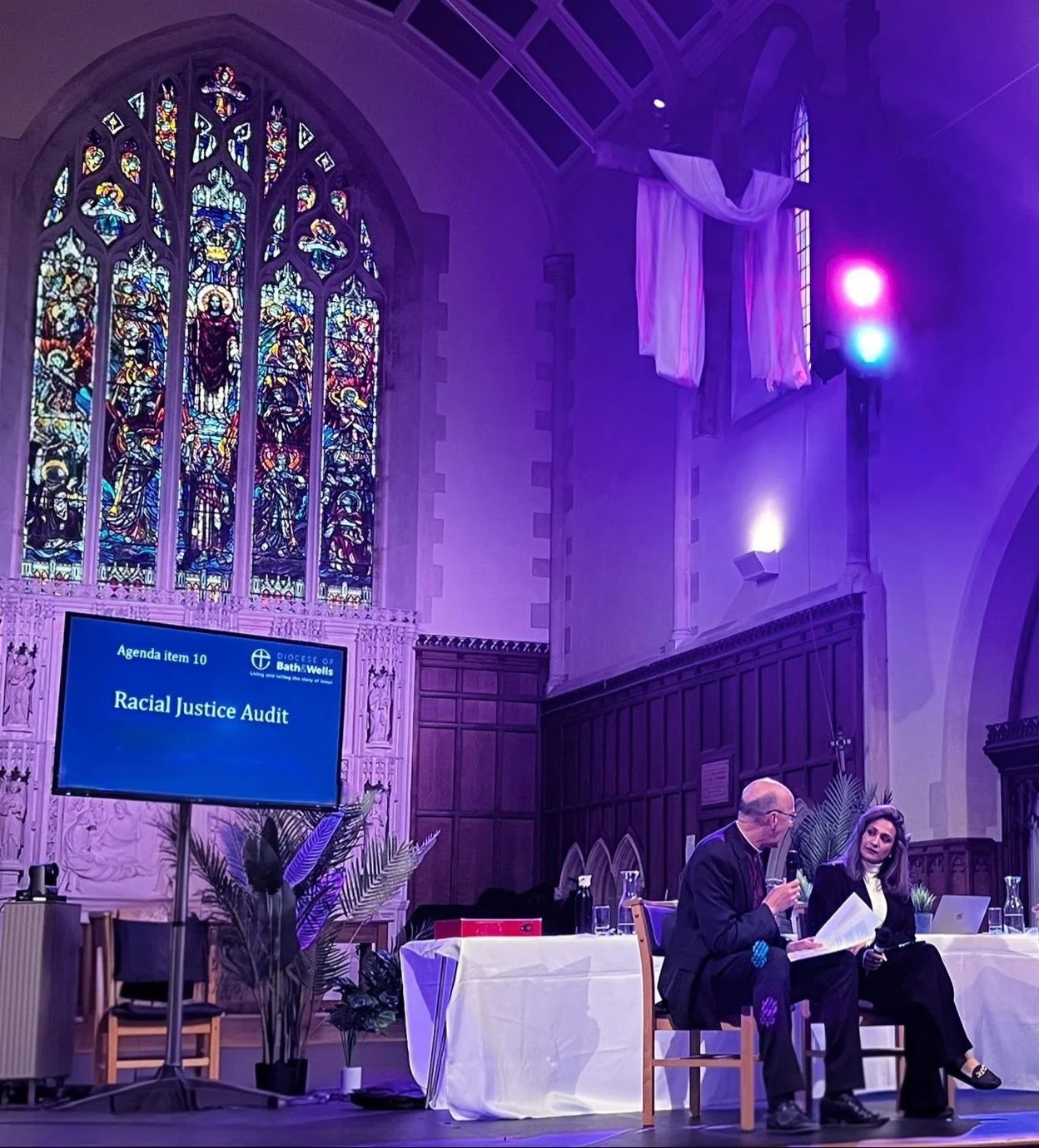 Anira Khokhar speaking on a church stage beside a screen reading “Agenda item 10, Diocese of Bath & Wells, Racial Justice Audit”