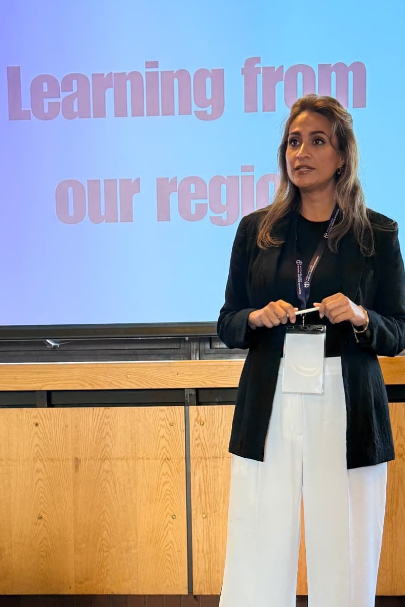 Anira Khokhar standing and speaking at a workshop, in front of her slide reading “Learning from our region”